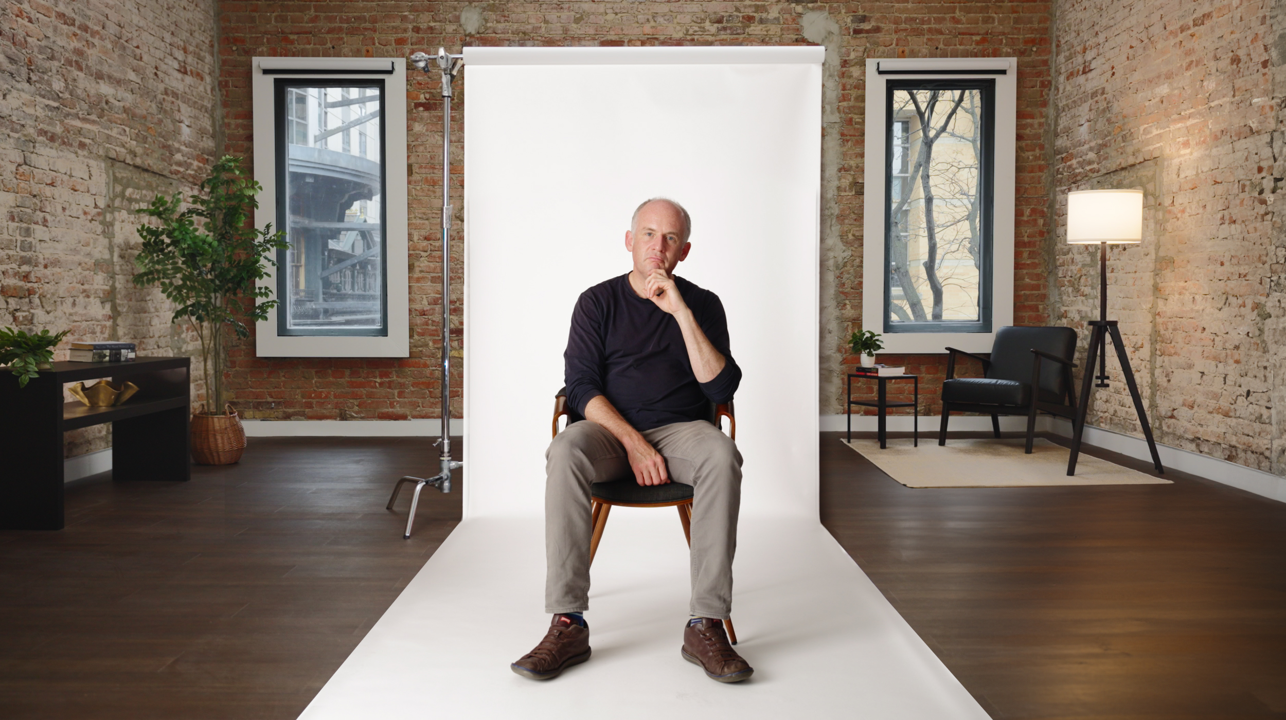 A person sitting on a chair in a cozy interior with brick walls, two windows, plants, and a lamp.