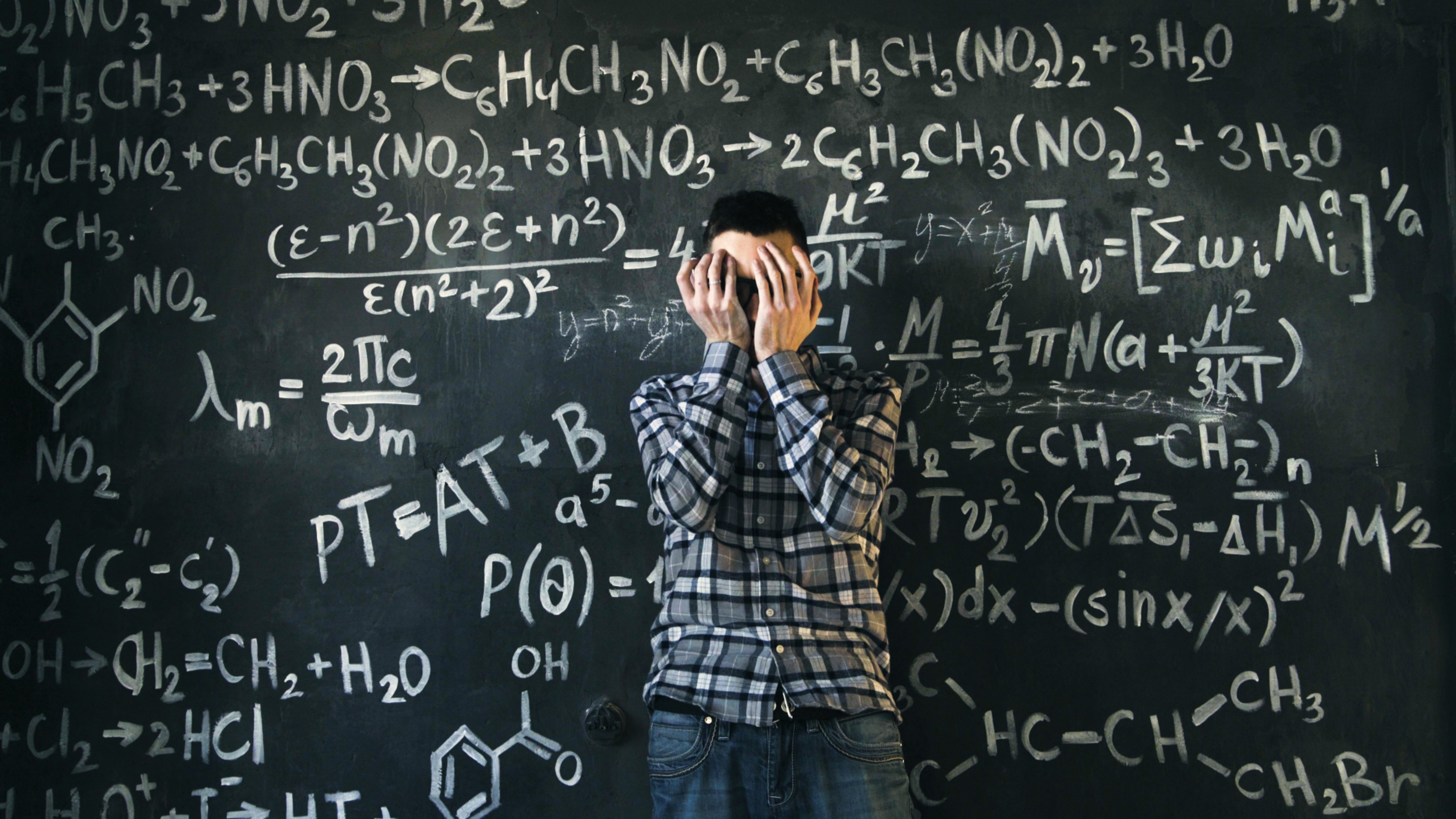 A person stands in front of a chalkboard filled with complex mathematical formulas, covering their face with their hands.