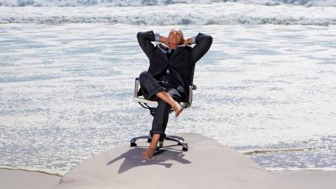 A man in a suit, embodying great management, relaxes on an office chair by the beach, with tranquil waves rolling in the background.