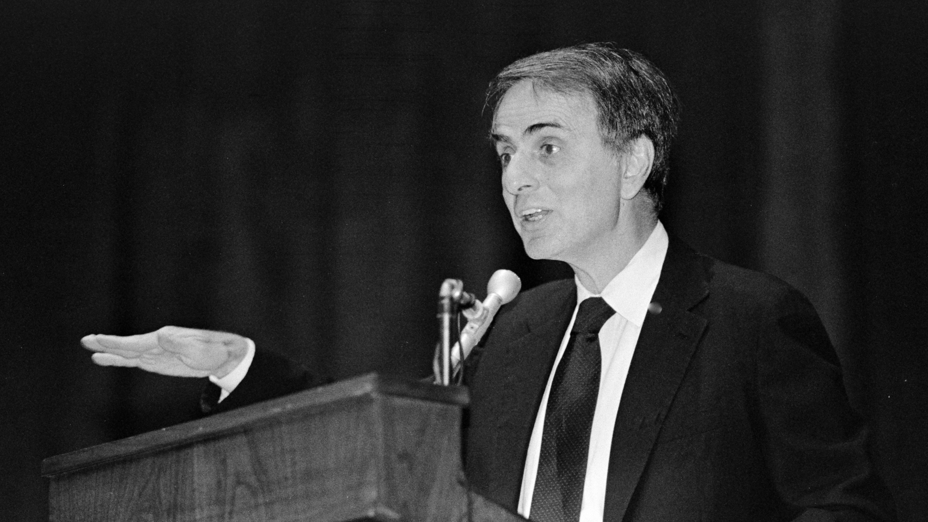 A man in a suit and tie speaks at a podium with a microphone, gesturing with his right hand.