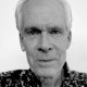 Black and white portrait of an older man with a neutral expression, wearing a patterned shirt, against a plain background.