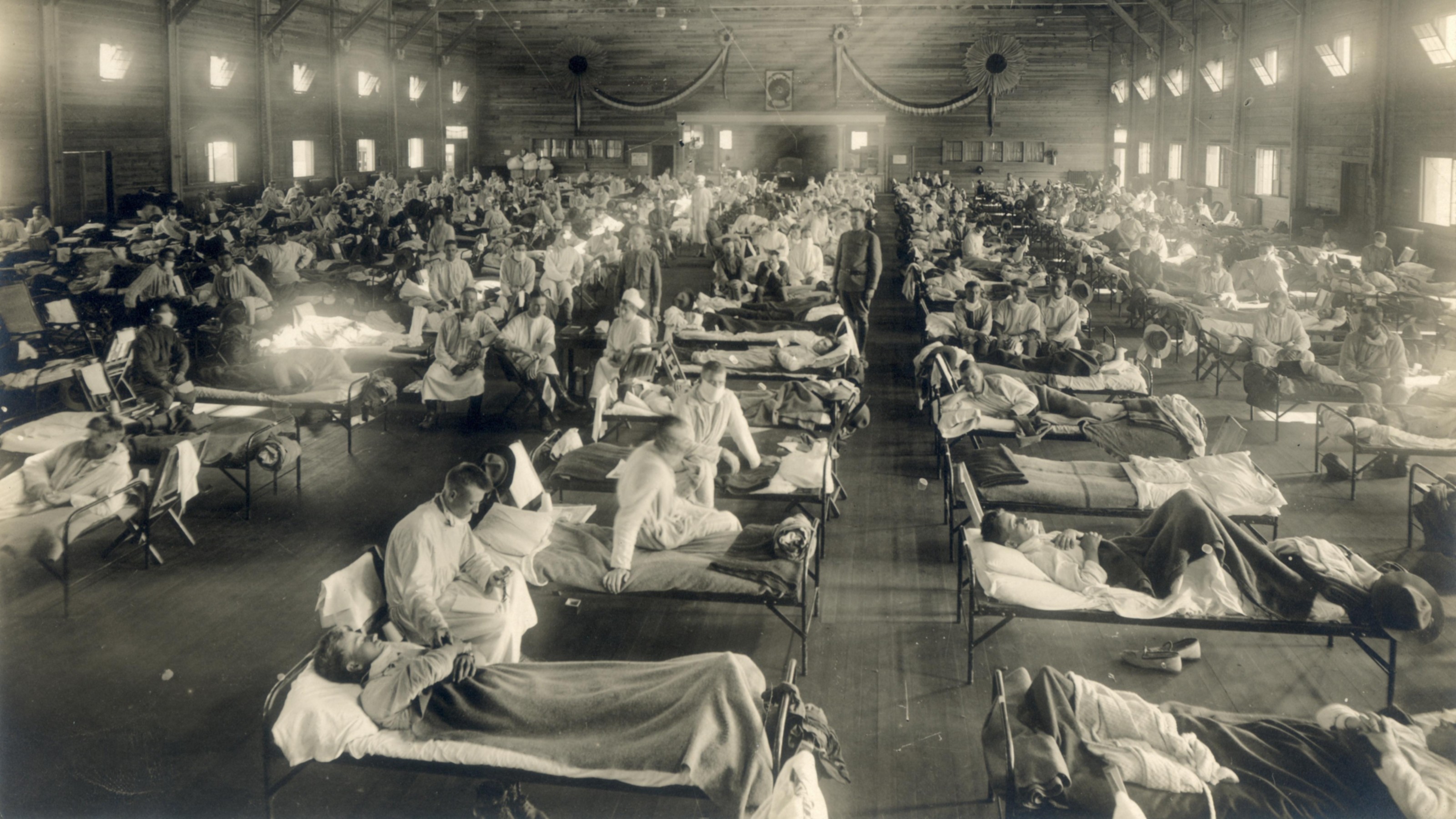 Large hall with rows of hospital beds occupied by patients and attended by medical staff, likely during an early 20th-century medical crisis.