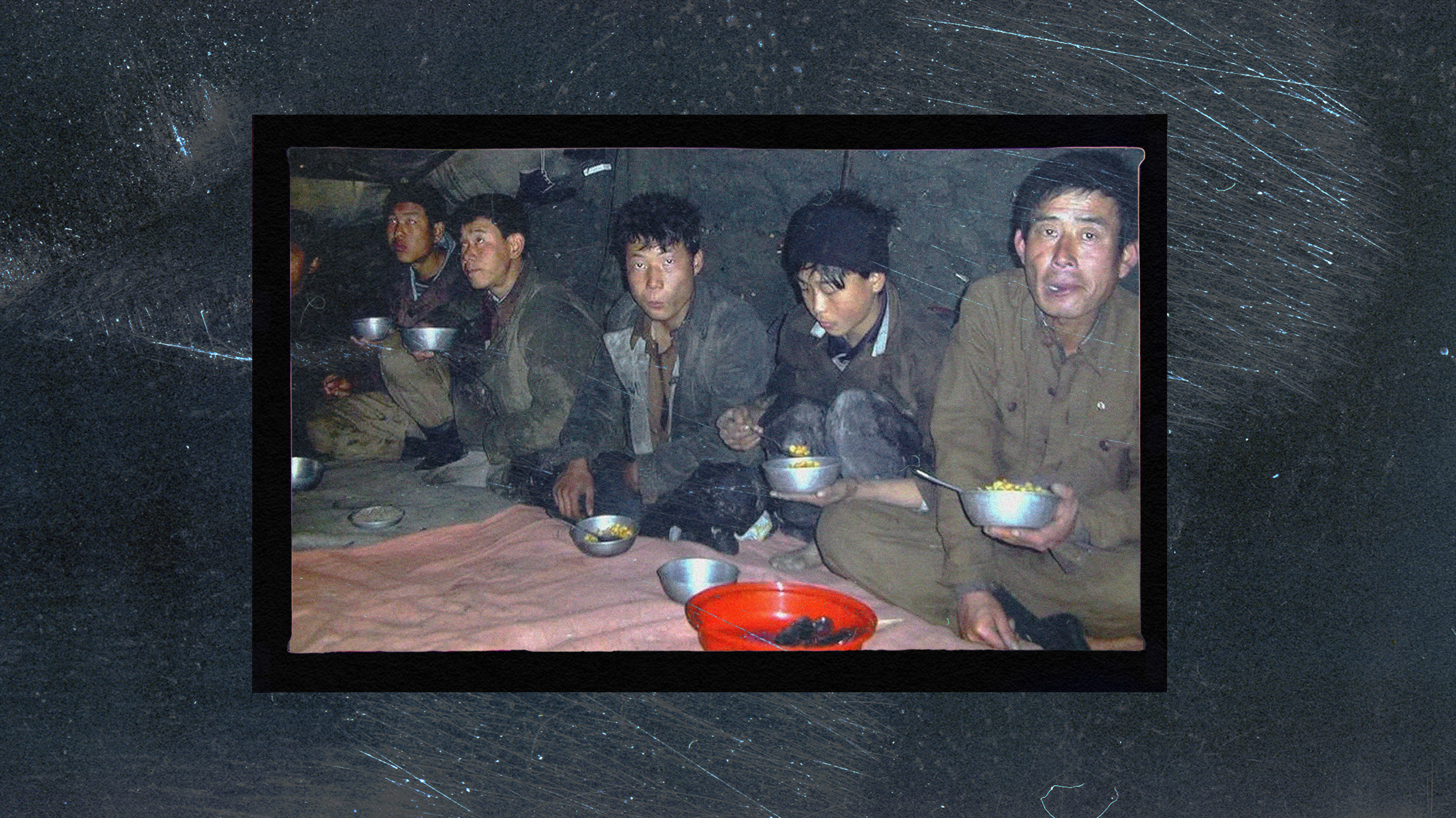 Five men, immersed in conversation, sit indoors on a blanket, eating from metal bowls. Despite the cloud of ignorance surrounding them about modern science, they share stories and laughter as a red bowl rests prominently at their center.