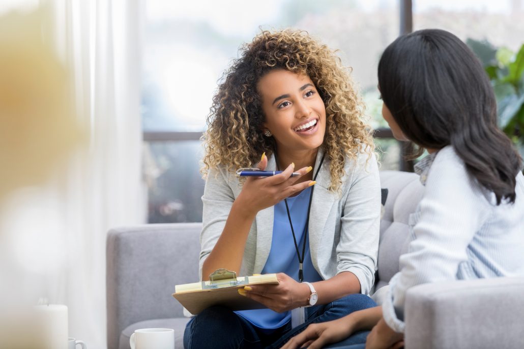 Positive young female therapist gestures as she talks with a female client. The therapist smiles warmly as she talks with the young woman.