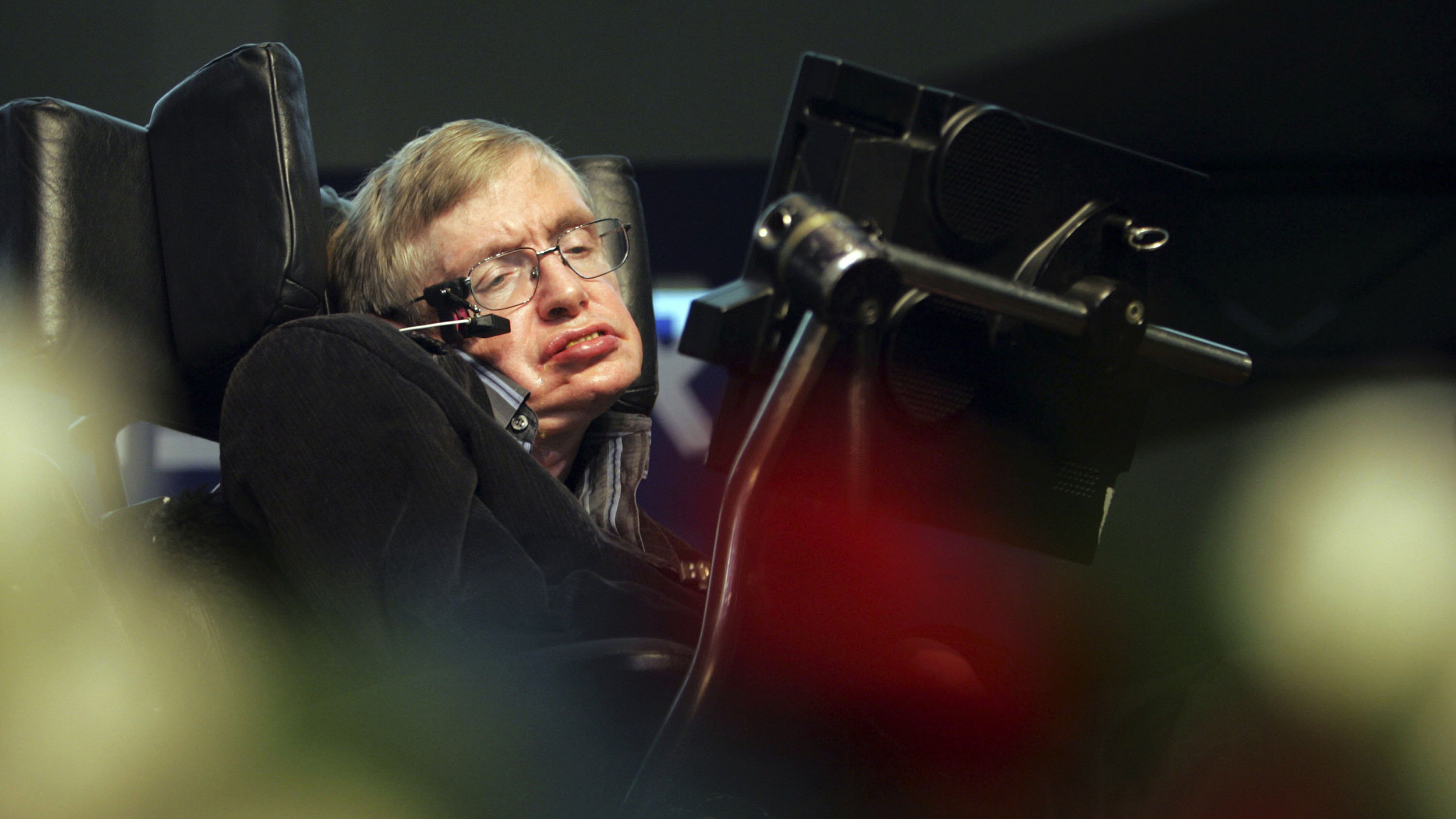 A person seated in a wheelchair uses a communication device with a Stephen Hawking-like voice, blurred flowers gently framing the scene in the foreground.