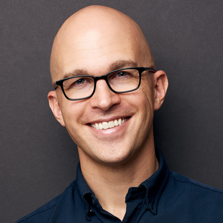 A smiling person with glasses and a shaved head is wearing a dark collared shirt against a dark background.