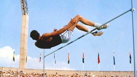 An athlete skillfully executes the Fosbury Flop, soaring gracefully in mid-air over the high jump bar, while colorful flags flutter and spectators watch in awe.