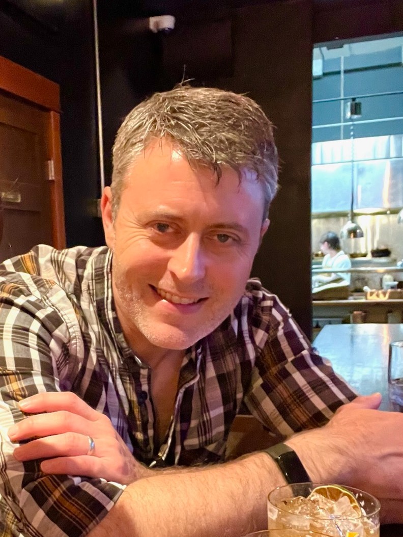 A man with short gray hair and wearing a plaid shirt is seated at a wooden table in a dimly lit restaurant, smiling at the camera.