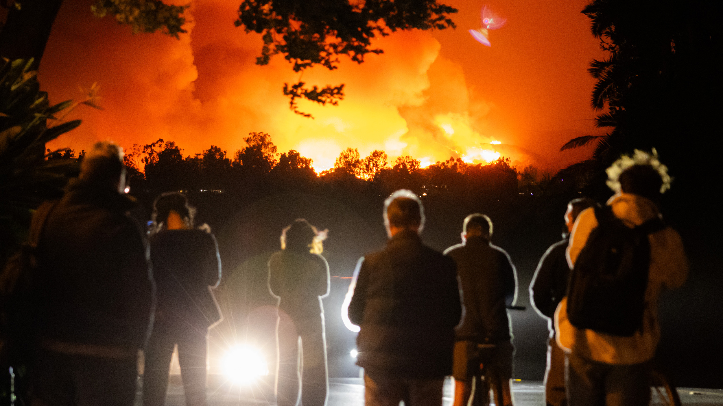 A group of people watches a large wildfire at night, with flames lighting up the sky in the background.