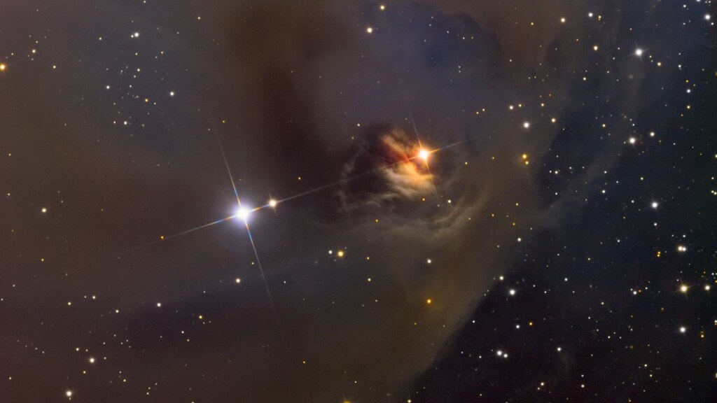 A dark nebula with dimming T Tauri stars nestled among dust and gas. The nebula casts a shadowy silhouette against a backdrop of numerous smaller stars in space.