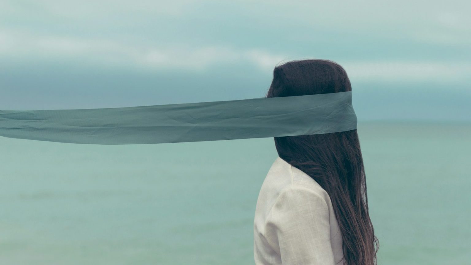 A person with long hair faces the ocean, wearing a white shirt. A sheer gray fabric is draped over their face, obscuring their eyes. The background is cloudy with a view of the sea.