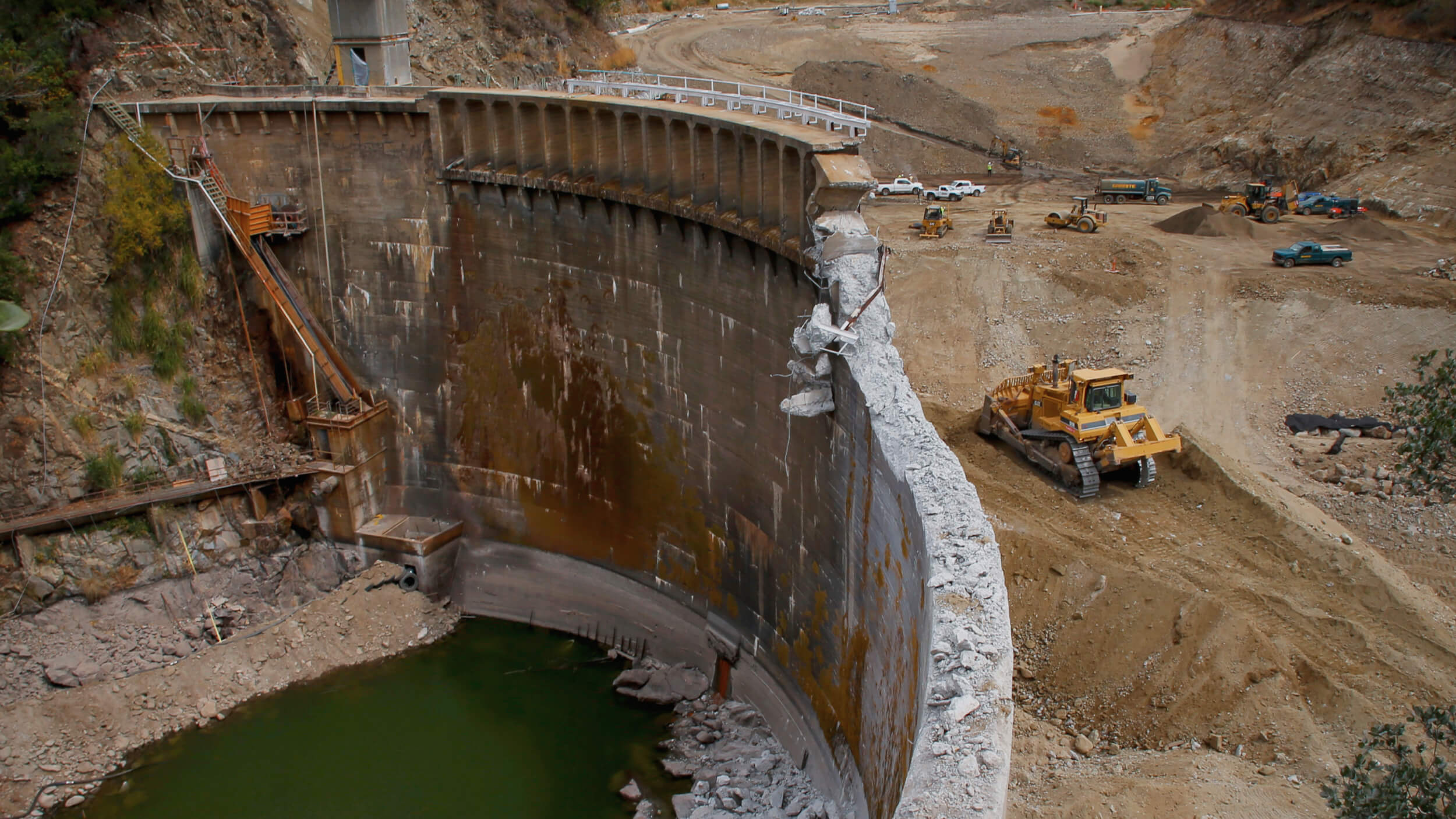 Construction equipment stands poised near a large, partially demolished dam wall, signaling the ongoing dam removal process beside a pool of green water.