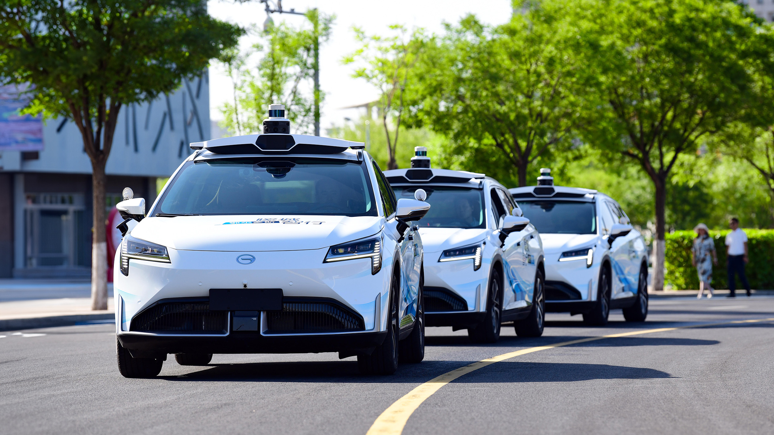 Three white autonomous cars drive down a city street lined with trees.