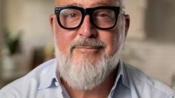 A person with a bald head, white beard, and black glasses is wearing a light blue shirt, standing in a softly lit kitchen.