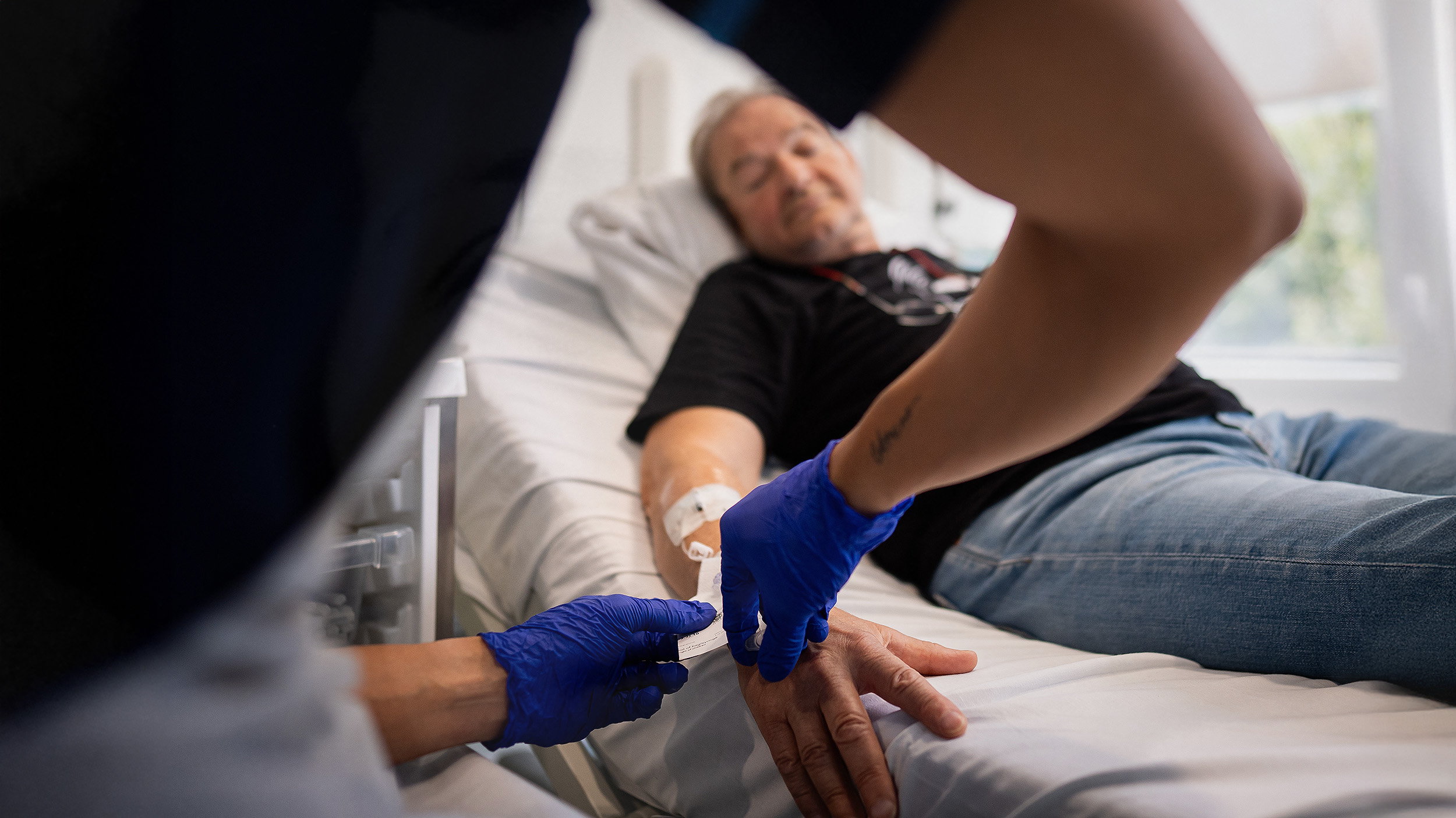 A healthcare worker wearing blue gloves gently inserts an IV into a man's hand as he lies on a hospital bed, battling lung cancer.